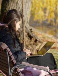 Woman in a dark coat sits on a park bench, typing on a laptop by a lake with autumn trees in the background.