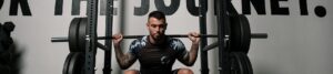 Tattooed man in a camouflage-patterned shirt squats in a power rack, lifting a heavily loaded barbell for a back squat in a gym.