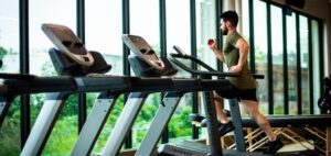 Man in a green shirt running on a treadmill in a bright gym with large windows.