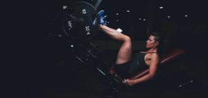 Woman in a black athletic outfit on a leg press machine, pushing a heavy weight in a dimly lit gym.