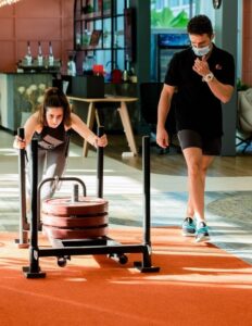 Woman pushes a loaded weight sled on an orange gym mat as a masked trainer watches indoors.