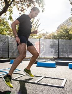 Young man in a black T-shirt and shorts jumping over an agility ladder in an outdoor gym setting, sunny day.