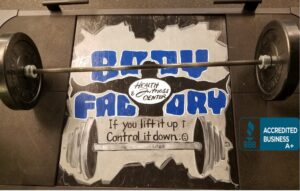 Barbell with weight plates on a gym platform; a torn poster behind reads 'Health & Fitness Center' with a motivational slogan about lifting and lowering the weight.