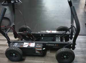 Low-angle view of a black four-wheeled pedal go-kart frame with thick tires and exposed wires on a wooden floor